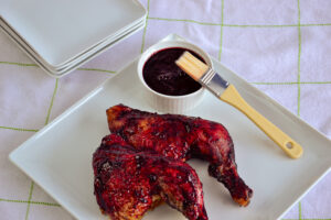 Grilled chicken leg resting on a plate, brushed with haskap BBQ sauce, with a ramekin of sauce nearby.
